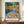 May include: Colorful Matisse exhibition poster in thin brown frame on wooden sideboard against cream walls with decorative molding. Artwork shows vibrant Fauve-style landscape with figures by water in blues, oranges, yellows, and greens. White ceramic vase and stacked books nearby provide scale reference.