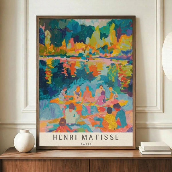 May include: Colorful Matisse exhibition poster in thin brown frame on wooden sideboard against cream walls with decorative molding. Artwork shows vibrant Fauve-style landscape with figures by water in blues, oranges, yellows, and greens. White ceramic vase and stacked books nearby provide scale reference.
