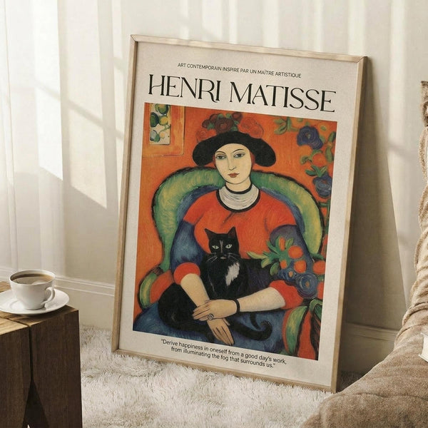 May include: Framed Henri Matisse poster showing a woman in red dress holding a black and white cat, seated in green chair against orange floral background. Light wood frame leaning against white wall corner on fluffy white rug, with coffee cup on wooden block nearby and beige blanket visible.
