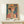 May include: Framed art print featuring a woman in an orange straw hat with geometric patterned background in vibrant blues, oranges, and greens. The colorful poster displays in a light wooden frame on a white wooden shelf alongside a small wrapped gift with pine sprig, against a cream wall in natural lighting.