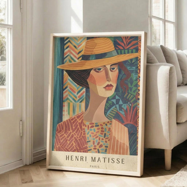May include: Large framed art print featuring a woman wearing an orange wide-brimmed hat in colorful Art Deco style with geometric patterns in teal, coral, and green tones. The print shows "Henri Matisse Paris" text at bottom and is displayed in a light wooden frame leaning against white walls near a beige upholstered chair in a bright, modern living room with large windows.