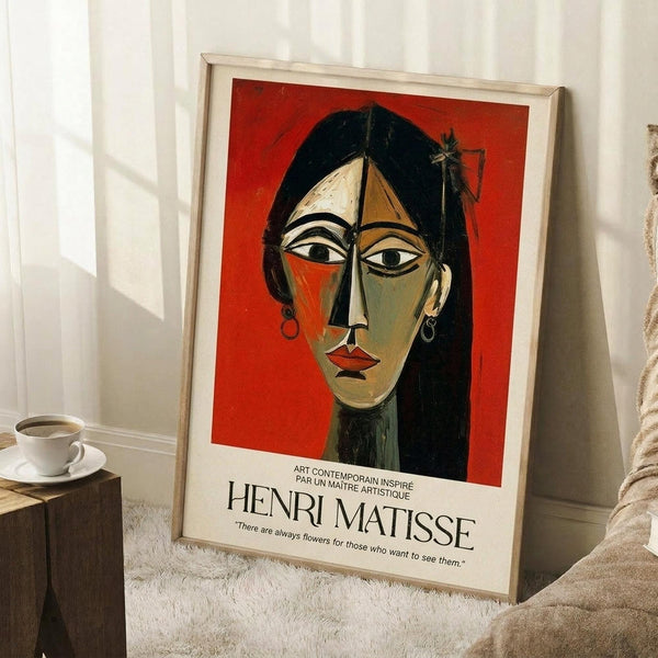 May include: Framed poster featuring a cubist-style portrait of a woman with geometric facial features, black hair, and earrings against a vibrant red background. The artwork shows angular, stylized proportions with bold colors including orange, green, and cream tones. French text appears below the image with an inspirational quote. The thin wooden frame leans against white paneled walls on a textured cream rug, with a coffee cup on a wooden block nearby.