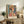 May include: Framed colorful portrait poster of woman wearing orange sun hat, featuring geometric patterns and vibrant blues, oranges, and greens in artistic style. Display shows poster leaning against white wall on rustic wooden console table beside fern plant, books, ceramic vase, and cream upholstered chair with brown leather pillow in cozy living room setting.