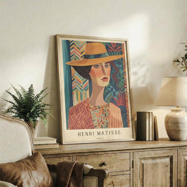 May include: Framed colorful portrait poster of woman wearing orange sun hat, featuring geometric patterns and vibrant blues, oranges, and greens in artistic style. Display shows poster leaning against white wall on rustic wooden console table beside fern plant, books, ceramic vase, and cream upholstered chair with brown leather pillow in cozy living room setting.