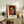May include: Framed Henri Matisse-style portrait poster featuring a woman's face in cubist style with red background, displayed on rustic wooden sideboard alongside fern plant, ceramic vase, and stacked books in warm neutral living room setting with beige armchair visible.