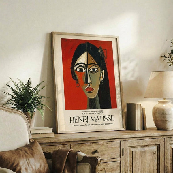 May include: Framed Henri Matisse-style portrait poster featuring a woman's face in cubist style with red background, displayed on rustic wooden sideboard alongside fern plant, ceramic vase, and stacked books in warm neutral living room setting with beige armchair visible.