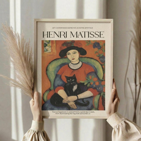 May include: Person in cream ruffled sleeves holding framed Henri Matisse art print featuring woman in black hat and red dress holding black and white cat, sitting in green armchair against orange floral background. Dried wheat stems visible on left. Warm natural lighting with soft white curtains in background.