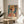 May include: A colorful art poster in a thin wooden frame propped on a round wooden side table next to a beige upholstered chair. The artwork depicts a woman wearing an orange wide-brimmed hat with geometric patterns and vibrant tropical motifs in the background. Soft natural light creates shadows on the white wall behind. A palm plant is partially visible on the left side.