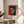 May include: Framed Henri Matisse exhibition poster featuring a cubist-style woman's portrait with geometric facial features in green, yellow and red tones against a bright red background. The cream-colored poster displays in a thin wooden frame, positioned on a round wooden side table next to a beige upholstered chair. Soft natural light creates shadows across white walls, with green plant leaves visible in the foreground.