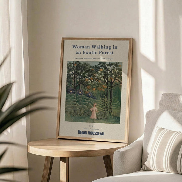 May include: A framed art print featuring Henri Rousseau's painting of a woman in white dress walking through a lush green jungle with dense tropical foliage and trees. The poster has cream background with blue text reading "Woman Walking in an Exotic Forest" and artist name. Displayed on a round wooden side table next to a beige upholstered chair with striped pillow, with a plant visible in soft natural lighting.
