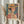 May include: Hands holding a framed art print featuring a woman wearing an orange wide-brimmed hat in colorful geometric Matisse-inspired style with patterned background of chevrons and tropical motifs. Person in beige ruffled sleeves holds dried pampas grass stems beside the artwork against white curtained window backdrop.