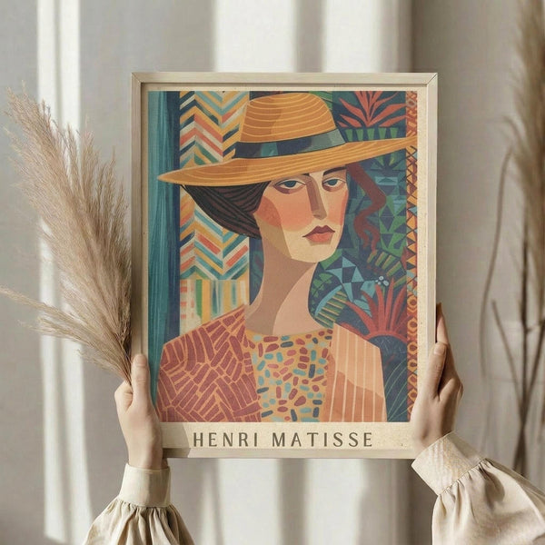 May include: Hands holding a framed art print featuring a woman wearing an orange wide-brimmed hat in colorful geometric Matisse-inspired style with patterned background of chevrons and tropical motifs. Person in beige ruffled sleeves holds dried pampas grass stems beside the artwork against white curtained window backdrop.