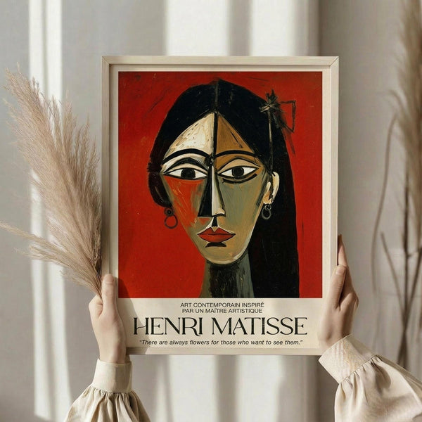 May include: Person in cream blouse holding framed art print against white curtained window, with dried pampas grass visible. The print features a stylized portrait of a woman with black hair and geometric facial features in green, orange, and red tones against a red background. White frame with text below the artwork including Henri Matisse attribution and French text.