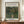 May include: Framed vintage art poster showing Henri Rousseau's exotic forest painting with lush green jungle foliage and a small figure in pink dress walking through dense tropical plants. The cream-colored poster has blue text reading "Woman Walking in an Exotic Forest" and displays on a wooden credenza next to a white ceramic vase and stacked books in a bright interior room with white paneled walls.