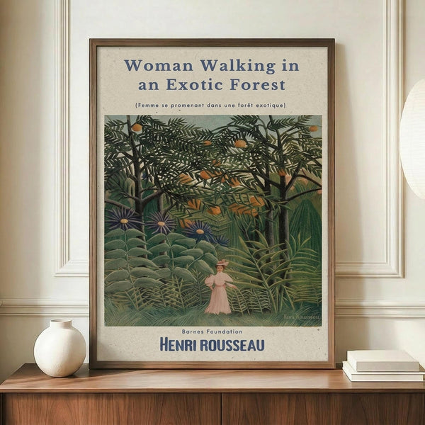 May include: Framed vintage art poster showing Henri Rousseau's exotic forest painting with lush green jungle foliage and a small figure in pink dress walking through dense tropical plants. The cream-colored poster has blue text reading "Woman Walking in an Exotic Forest" and displays on a wooden credenza next to a white ceramic vase and stacked books in a bright interior room with white paneled walls.