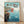 May include: Framed Studio Ghibli Ponyo movie poster in natural wood frame leaning against white wall on light wooden floor. The artwork shows red-haired Ponyo in orange dress jumping from rocky coastal cliffs into bright blue ocean water with splashing waves. Japanese text and "PONYO" appear at top against soft blue-green sky. Classic anime illustration style with warm earth tones contrasting against cool ocean blues.