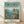 May include: Framed poster print in light wood frame leaning against white wall on wooden floor. Features Studio Ghibli's Ponyo movie poster with young girl in red dress standing on green cliff overlooking blue ocean and coastal village. Japanese and English text at top, Studio Ghibli credits at bottom. Soft natural lighting creates gentle shadows on textured white wall.