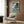 May include: Framed Studio Ghibli Ponyo poster displayed on wood-paneled wall above cream upholstered armchair with geometric pillow. Artwork shows red-haired character in wooden boat on choppy ocean waves near forested cliffs under stormy sky. Modern reading nook with brass floor lamp, side table with open book, and natural lighting from window with curtains.