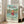 May include: Framed Studio Ghibli Ponyo poster displayed in bright living room with beige sofa and white walls. Artwork shows red-haired child character running through ocean waves with Japanese coastal village in background. Turquoise water, green hills, and traditional buildings create colorful anime-style illustration in light wood frame on wooden floor.