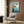May include: Studio Ghibli Ponyo movie poster mounted on wood-paneled wall above cream upholstered armchair with geometric pillow. Poster shows red-haired child jumping from seaside cliff into blue ocean water. Modern reading nook with brass floor lamp, open book on dark side table, and natural lighting from window with sheer curtains.