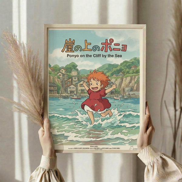 May include: Hands holding a framed Studio Ghibli Ponyo poster featuring red-haired Ponyo running through ocean waves toward a coastal village. The anime-style artwork shows turquoise water, green hills, and traditional buildings under a cloudy sky. Light wooden frame against white curtains with dried pampas grass visible on the left side.