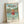 May include: Framed Studio Ghibli Ponyo movie poster in light wood frame leaning against white wall. Features young red-haired Ponyo in red dress running through ocean waves with coastal Japanese village and green hills in background. Bright anime art style with blue-green water, cloudy sky, and Japanese text above English title.