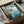 May include: Framed Studio Ghibli Ponyo poster showing red-haired character running across blue ocean waves near rocky coastal cliffs, packaged in cardboard shipping box with protective wrapping and wooden frame corners visible, photographed on white surface with partial view of dark objects in background