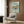 May include: Studio Ghibli Ponyo movie poster in light wood frame hanging on warm brown wood paneled wall above cream upholstered armchair with geometric pillow. Poster shows red-haired child character running through ocean waves with Japanese and English text at top. Modern reading nook setting with brass floor lamp and side table with open book.
