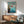 May include: Framed Studio Ghibli Ponyo poster showing red-haired character jumping over ocean cliff, displayed above wooden sideboard with white ceramic lamp and mid-century modern teal armchair in sunlit room with white walls