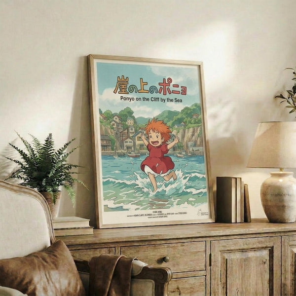 May include: Framed Studio Ghibli Ponyo poster displayed on rustic wooden console table in cozy living room setting. Artwork shows red-haired child running through ocean waves with Japanese coastal town background. Natural wood frame, surrounded by fern plant, books, ceramic vase, and beige upholstered furniture with brown leather cushions.