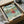 May include: Framed Studio Ghibli Ponyo poster in black frame, packaged in cardboard box with tissue paper. Artwork shows red-haired boy Ponyo running through ocean waves with coastal town and green cliffs in background. Japanese text at top reads "Ponyo on the Cliff by the Sea." Colorful anime illustration with turquoise water, traditional buildings, and lush vegetation. Frame sits in shipping materials on white surface.