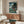 May include: Framed Studio Ghibli Ponyo poster displayed above a mid-century modern walnut credenza in a minimalist living room. The colorful artwork shows a red-haired character in a wooden boat on stormy ocean waves near a cliff with houses. White sculptural chair and round glass coffee table complete the Scandinavian-style interior with cream walls and natural lighting.