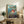 May include: Framed Studio Ghibli Ponyo movie poster displayed on rustic wooden sideboard in cozy living room setting. Poster shows red-haired Ponyo character swimming in ocean waves with coastal town background. Natural wood frame, surrounded by fern plant, ceramic vases, stacked books, and neutral armchair with brown throw pillow.