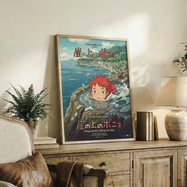 May include: Framed Studio Ghibli Ponyo movie poster displayed on rustic wooden sideboard in cozy living room setting. Poster shows red-haired Ponyo character swimming in ocean waves with coastal town background. Natural wood frame, surrounded by fern plant, ceramic vases, stacked books, and neutral armchair with brown throw pillow.