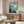 May include: Studio Ghibli Ponyo movie poster mounted on white wall above wooden sideboard in modern living room. Poster shows red-haired child jumping into blue ocean water from rocky cliff. Mid-century furniture includes walnut credenza, white curved chair, oval glass coffee table, and minimalist table lamps with warm lighting.