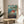 May include: Framed Studio Ghibli Ponyo movie poster featuring red-haired character swimming in ocean waves with coastal village backdrop, displayed on round wooden side table next to beige upholstered chair with throw pillow, plant leaves visible in corner, natural lighting creating soft shadows on neutral wall.