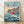 May include: Framed Studio Ghibli Ponyo movie poster featuring red-haired girl in red dress jumping over ocean waves with coastal village on cliffs in background. Wooden frame displayed against white wall on light wood floor with natural lighting creating soft shadows.