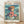 May include: Framed Studio Ghibli Ponyo movie poster in light wood frame leaning against white wall on wooden floor. Features anime-style illustration of young girl with flowing brown hair in red and blue dress sitting by turbulent ocean waves near rocky cliff. Soft natural lighting creates shadows on white textured wall. Poster shows Japanese text at top with English translation, ocean scene in blues and greens, and film credits at bottom.