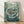 May include: Framed poster print in light wood frame leaning against white wall on wooden floor. Artwork shows underwater scene with four children floating together surrounded by swirling water, colorful fish, and jellyfish in teal and turquoise tones. White and Japanese text at bottom reads "PONYO" and "PONYO ON THE CLIFF BY THE SEA" with Studio Ghibli credits. Soft natural lighting creates gentle shadows on textured white wall.