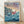 May include: Framed Studio Ghibli Ponyo movie poster print leaning against white wall on wooden floor. Shows red-haired character swimming in blue ocean waves with fish, clifftop village houses above, sunset sky. Japanese and English text at top. Natural wood frame, soft window light casting shadows on textured white wall.