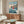 May include: Studio Ghibli Ponyo poster mockup in modern living room setting with wooden sideboard, white sculptural chair, and glass coffee table. Poster shows red-haired child character Ponyo running on ocean waves with coastal village background in blue, pink and coral tones. Thin wooden frame against cream walls with natural lighting.