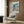 May include: Framed Studio Ghibli Ponyo movie poster mounted on wood-paneled wall above beige upholstered armchair with geometric patterned pillow. Poster shows young red-haired character by ocean waves in anime art style with Japanese and English text. Modern living room setting with brass floor lamp, side table with open book, and natural lighting from window with curtains.