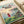 May include: Studio Ghibli-style poster print displayed on white bedding next to a cream pillow. The artwork shows a young girl with red hair in a red dress standing on a grassy cliff overlooking a coastal village with traditional Japanese houses and fishing boats in the harbor. Features soft watercolor-style illustration with pastel sky colors and Japanese text at the top along with "PONYO A FILM BY HAYAO MIYAZAKI" in English lettering.