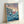 May include: Framed poster print of Studio Ghibli's Ponyo displayed against a white wall. The artwork shows the red-haired character Ponyo swimming in turbulent blue ocean waves with a clifftop village in the background under a purple and orange sunset sky. Japanese and English text appears at the top. The print is mounted in a light wooden frame and leans against the wall on what appears to be a white surface.