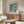 May include: Framed Studio Ghibli Ponyo poster mounted on white wall above mid-century walnut credenza in modern living room. Poster shows red-haired boy character against ocean backdrop with Japanese and English text. Room features cream oval chair, glass coffee table, white dome lamp, and wooden floor lamp with natural lighting.