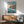May include: Framed Studio Ghibli Ponyo poster mounted on white wall above wooden sideboard with ceramic lamp. Features animated red-haired girl character running on ocean waves with coastal village in background. Mid-century modern living room setting with teal upholstered armchair, round coffee table, and natural sunlight streaming across scene.