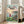 May include: Framed poster of animated film featuring young girl with red hair in red dress standing on coastal cliff overlooking seaside village with traditional houses, boats in harbor, and sunset sky. Poster shows Japanese text at top with "PONYO" and director credit below. Light wood frame leaning against beige sofa in bright living room with white walls and windows.
