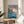 May include: Framed Studio Ghibli Ponyo movie poster displayed on wooden round side table next to beige upholstered chair. Poster shows red-haired girl character swimming in blue ocean waves with clifftop village in background, Japanese and English text at top. Natural lighting creates soft shadows on white wall behind minimal modern furniture setup with green plant visible.