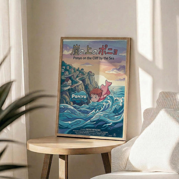 May include: Framed Studio Ghibli Ponyo movie poster displayed on wooden round side table next to beige upholstered chair. Poster shows red-haired girl character swimming in blue ocean waves with clifftop village in background, Japanese and English text at top. Natural lighting creates soft shadows on white wall behind minimal modern furniture setup with green plant visible.