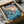 May include: Framed poster print in cardboard shipping box with protective packaging. Artwork shows anime-style character with red hair swimming in blue ocean waves near coastal cliff village. Japanese text at top with English subtitle "Ponyo on the Cliff by the Sea." Colorful illustration features sunset sky, traditional houses on rocky coastline, and dynamic water effects. Black frame against white tissue paper and brown cardboard packaging.
